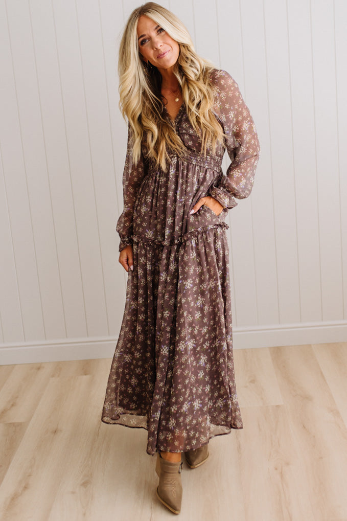 A woman modeling a long-sleeve, flowy maxi dress with a brown and beige floral pattern, standing in a room with light wood floors and white paneled walls.
