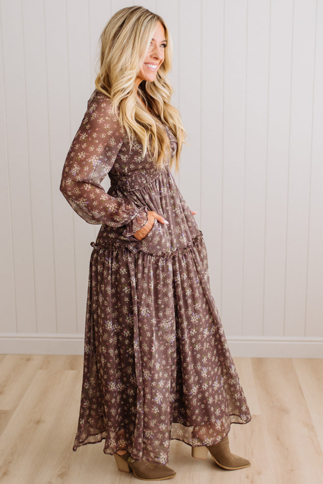 A woman modeling a long-sleeve brown maxi dress with a white floral pattern, standing in front of a white paneled wall and light wood floor.