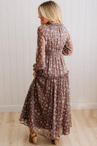 A woman wearing a long-sleeve, tiered maxi dress with a brown and beige floral pattern, shown from the back against a white paneled wall and light wood floor.