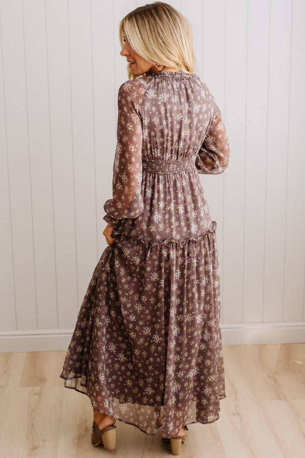 A woman wearing a long-sleeve, tiered maxi dress with a brown and beige floral pattern, shown from the back against a white paneled wall and light wood floor.
