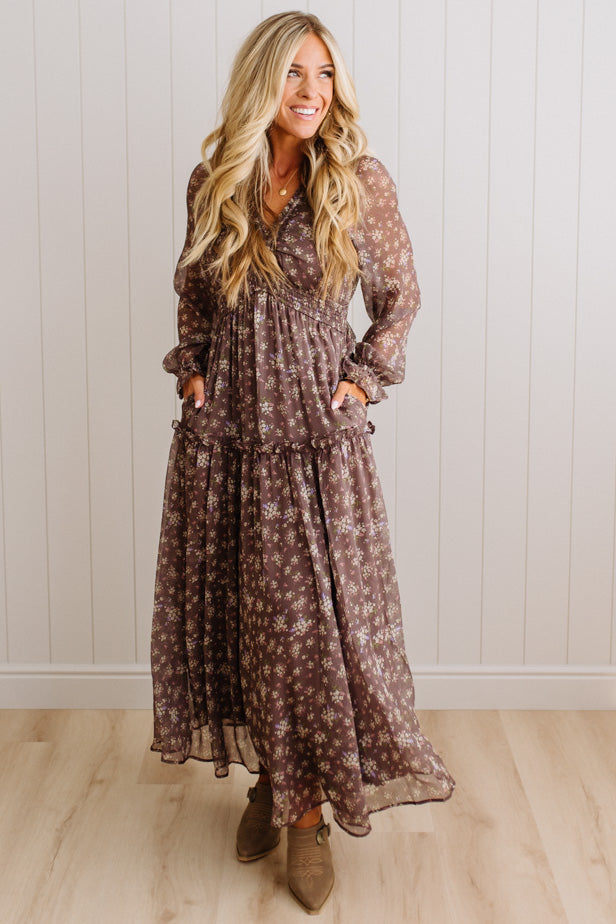 A woman modeling a long-sleeve, tiered maxi dress with a brown and beige floral pattern, standing in front of a white paneled wall and light wood floor.