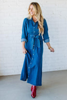 Woman wearing a blue denim dress with rolled-up sleeves and red boots against a white brick wall.