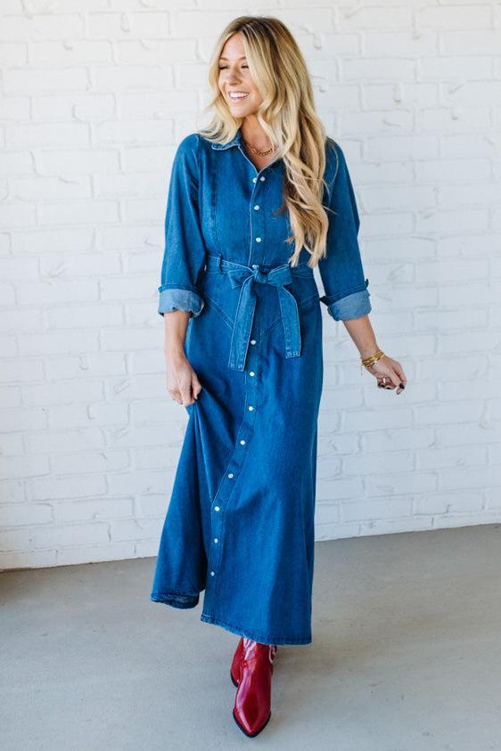 Woman wearing a blue denim dress with rolled-up sleeves and red boots against a white brick wall.