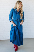 Woman wearing a blue denim dress with red shoes against a white brick wall.