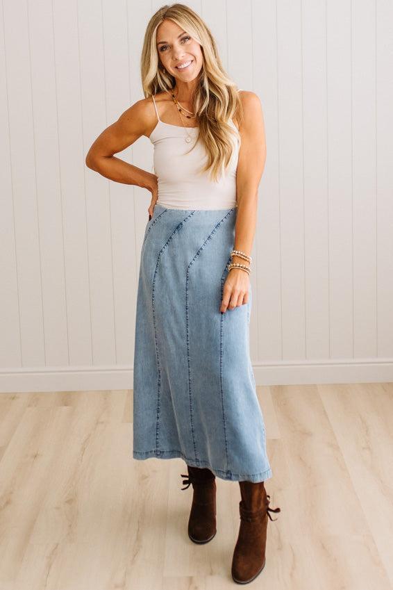 Blonde woman in a studio with light floor and a paneled wall behind, wearing a Denim Diagonal Line Detail Midi Skirt.