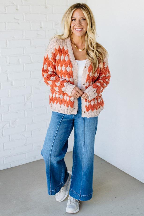 Woman wearing a colorful cardigan, white top, blue jeans, and sneakers against a white brick wall.