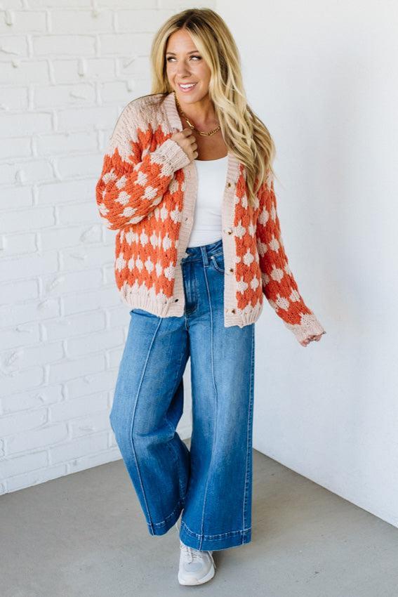 Woman wearing a colorful cardigan, white top, and blue jeans against a white brick wall.