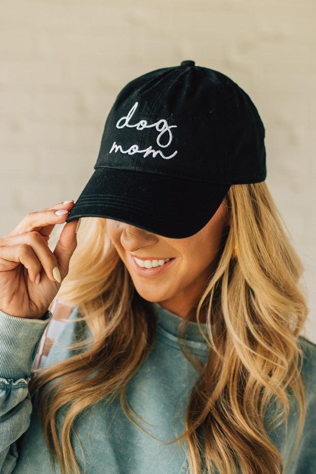 Blonde woman wearing a black hat with white cursive embroidery that say "dog mom" at the center front.