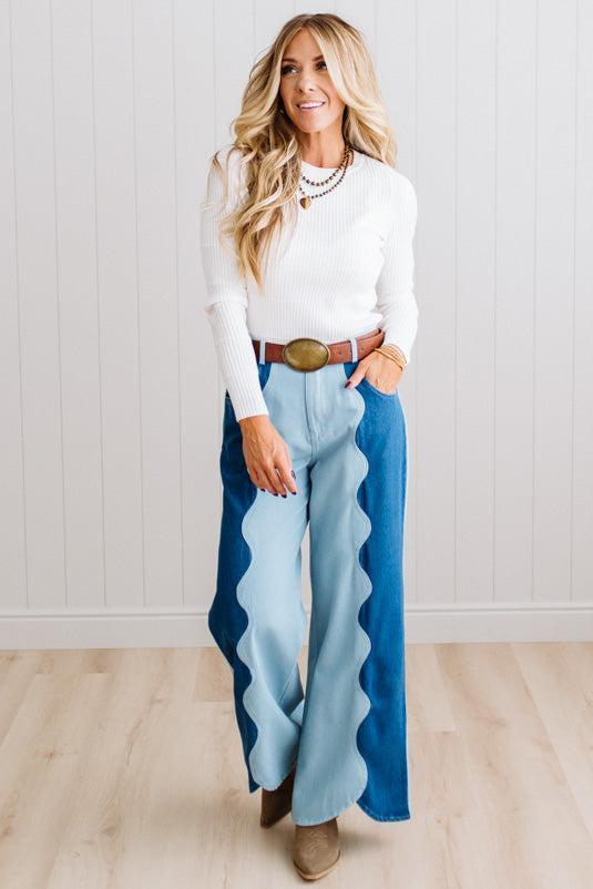 BLONDE WOMAN IN A BRIGHTLY LIT STUDIO WEARING A WHITE SWEATER, BROWN BELT AND SCALLOP WAVE DENIM OCEAN CURVE JEANS WITH TWO TONES.