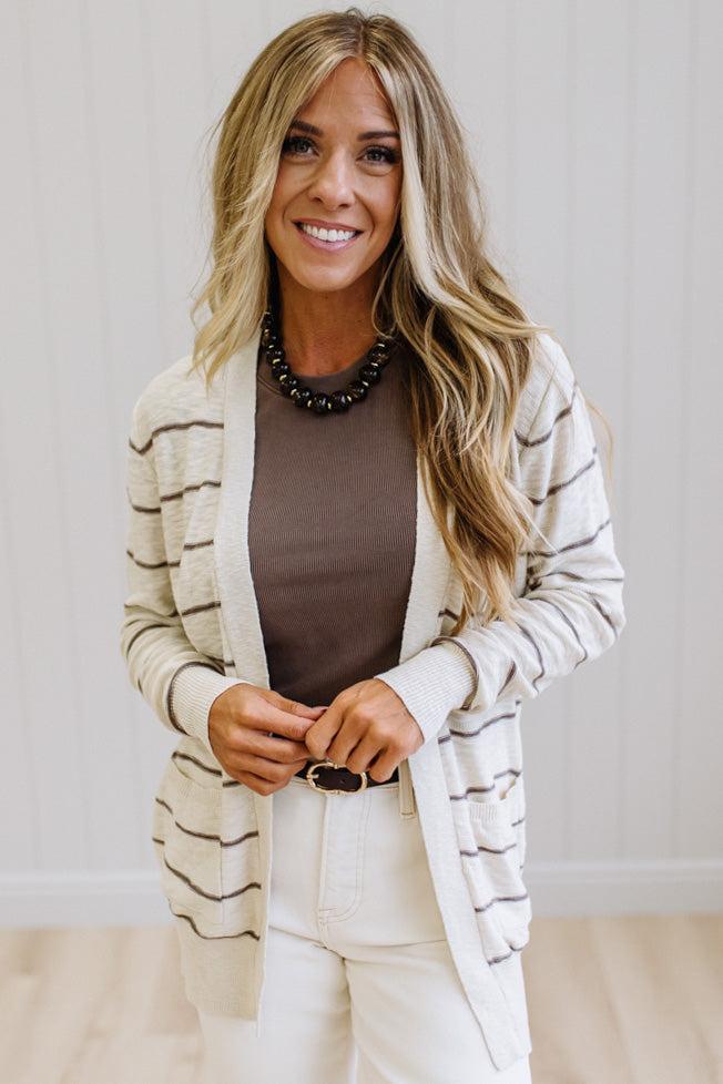 A smiling woman with blonde hair models a cream and brown striped cardigan over a brown top and white pants, standing against a white paneled wall.