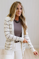 A woman modeling a cream and brown horizontally striped knit cardigan over a taupe top and white pants, standing against a light wood-paneled wall.