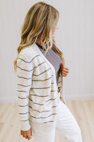 A woman wearing a cream knit cardigan with thin brown horizontal stripes over a mauve top and white pants, standing in a bright room with light wood floors and white paneled walls.