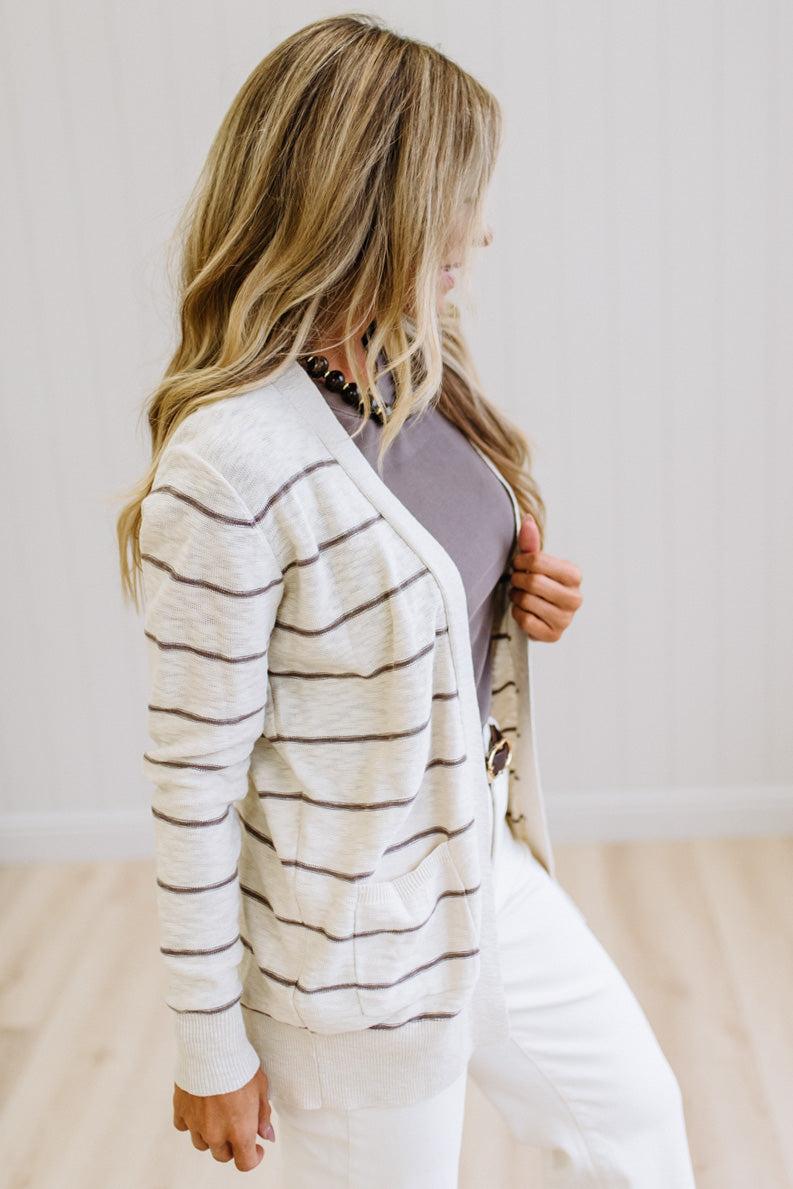 A woman wearing a cream knit cardigan with thin brown horizontal stripes over a mauve top and white pants, standing in a bright room with light wood floors and white paneled walls.