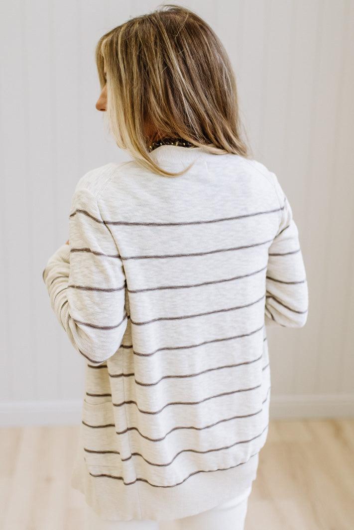 A woman viewed from behind wearing a cream-colored long-sleeve knit sweater with thin dark horizontal stripes, paired with white pants, set against a light wood-paneled wall and light wood floor.