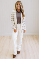A woman modeling a cream and brown striped knit cardigan over a taupe top and white wide-leg pants, standing in a bright room with light wood floors and white paneled walls.