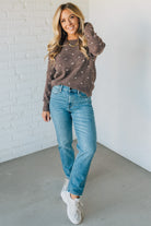 Woman wearing a brown sweater with white polka dots and blue jeans against a white brick wall.