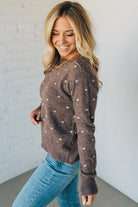 Woman wearing a brown sweater with white polka dots and blue jeans against a white wall.