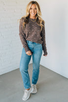 Woman wearing a brown polka dot sweater, blue jeans, and white sneakers against a white brick wall.