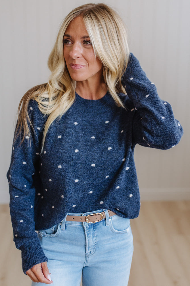 A woman wearing a navy blue knit sweater with small white polka dots, paired with light blue jeans and a tan belt, standing in a brightly lit indoor setting.