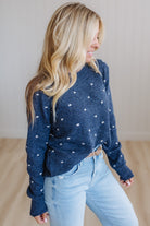 A woman wearing a navy blue knit sweater with small white polka dots and light blue jeans, standing in a brightly lit indoor room.