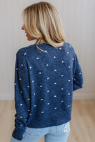 A woman with blonde hair is shown from behind wearing a navy blue knit sweater with small white polka dots, paired with light blue jeans, in an indoor setting with a light-colored wall and wooden floor.