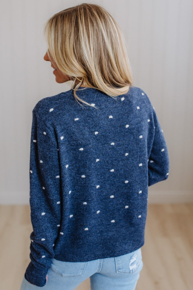 A woman with blonde hair is shown from behind wearing a navy blue knit sweater with small white polka dots, paired with light blue jeans, in an indoor setting with a light-colored wall and wooden floor.