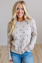 A smiling blonde woman wearing a light beige crewneck sweater with small black polka dots and blue jeans, standing in a brightly lit room with light wood floors and white paneled walls.
