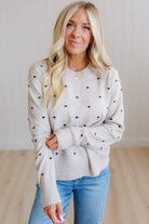 A woman with blonde hair wearing a light beige knit sweater with small black polka dots and blue jeans, standing in a brightly lit room with light wood floors.