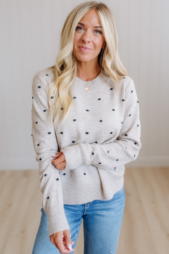 A woman with blonde hair wearing a light beige knit sweater with small black polka dots and blue jeans, standing in a brightly lit room with light wood floors.