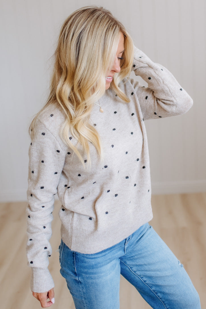 A woman wearing a light beige knit sweater with small black polka dots and blue jeans, standing in a brightly lit indoor room with light wood flooring.