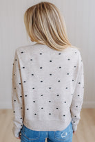 A woman viewed from behind wearing a light beige knit sweater with small black polka dots, paired with blue jeans, in a brightly lit indoor setting.