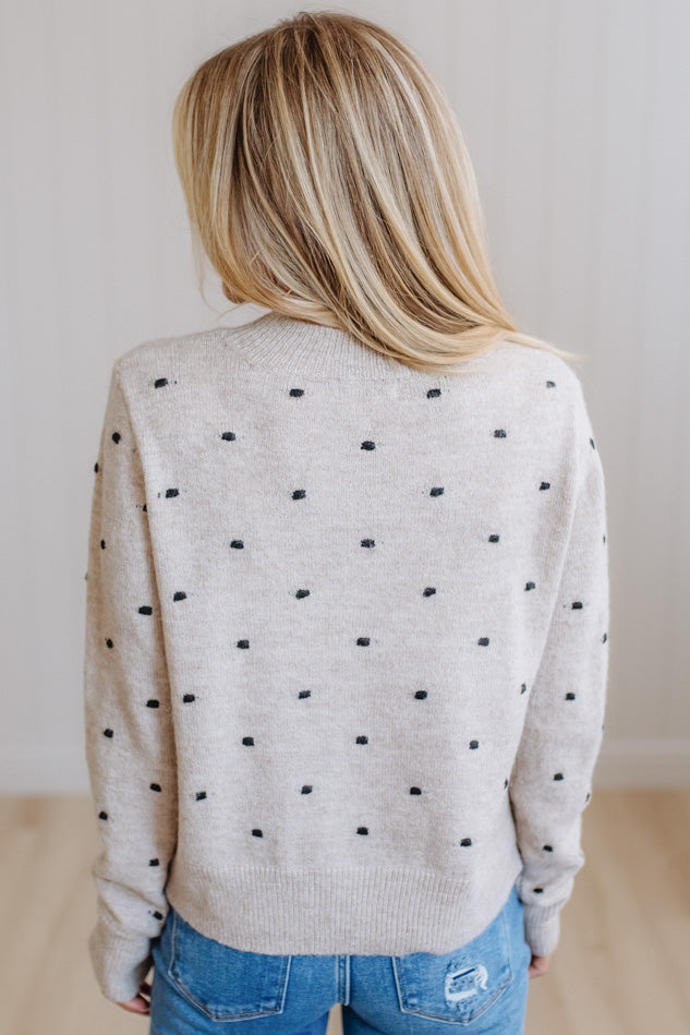 A woman viewed from behind wearing a light beige knit sweater with small black polka dots, paired with blue jeans, in a brightly lit indoor setting.