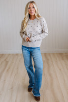 A woman modeling a light beige crewneck sweater with small black polka dots, paired with blue jeans and brown boots, standing in a room with light wood floors and white paneled walls.