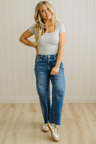 Woman wearing a striped shirt and blue jeans standing against a light wooden paneled wall.