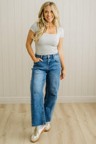 Woman wearing a white top and blue jeans standing against a light wooden paneled wall.
