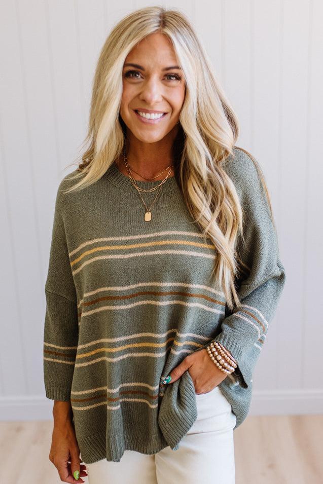A smiling blonde woman wearing a loose-fit knit sweater with horizontal stripes in olive green, tan, and brown, paired with white pants.