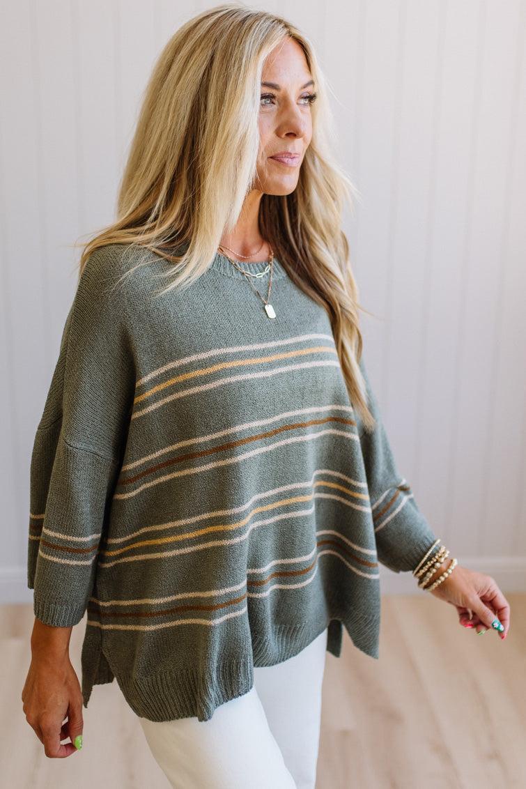 A woman wearing a muted sage green knit sweater with horizontal stripes in beige and mustard yellow, paired with white pants, standing against a light wood-paneled wall.