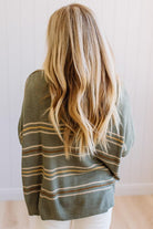 A woman viewed from behind wearing a loose olive green knit sweater with thin horizontal tan and white stripes, paired with white pants, set against a light-colored paneled wall.