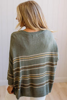 A woman viewed from behind wearing a loose knit sweater in muted sage green with horizontal stripes in tan, cream, and brown, set against a white paneled wall.