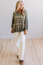 A woman modeling a loose-fitting knit sweater with horizontal stripes in olive green, beige, and brown, paired with off-white wide-leg pants and brown slide sandals, standing in a room with light wood floors and white paneled walls.