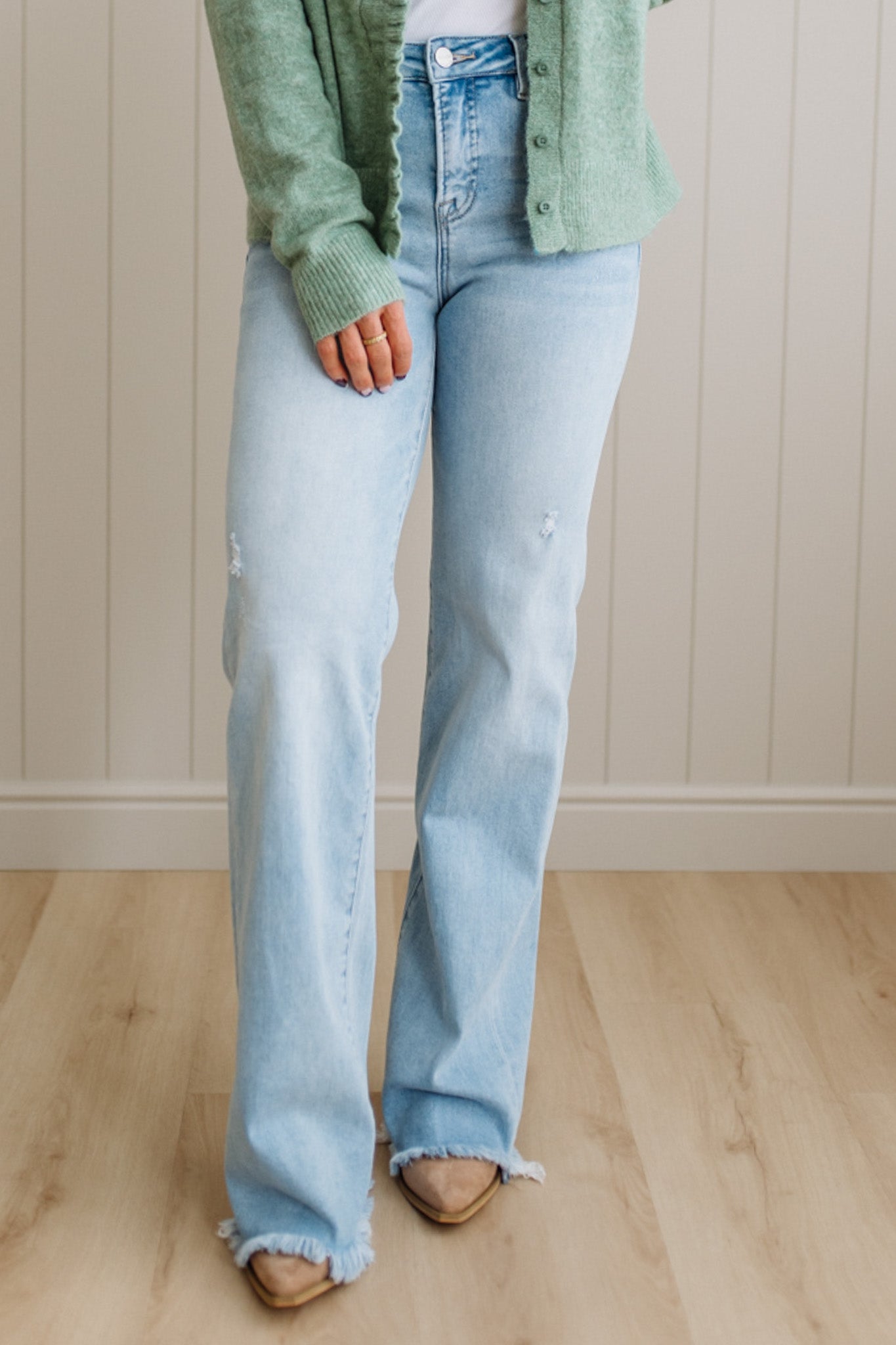 Person wearing light blue flared jeans and a green cardigan against a beige wall.