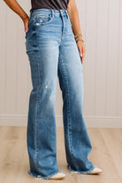 A person wearing light blue high-waisted flare jeans with a raw frayed hem, standing on a light wood floor against a white paneled wall.