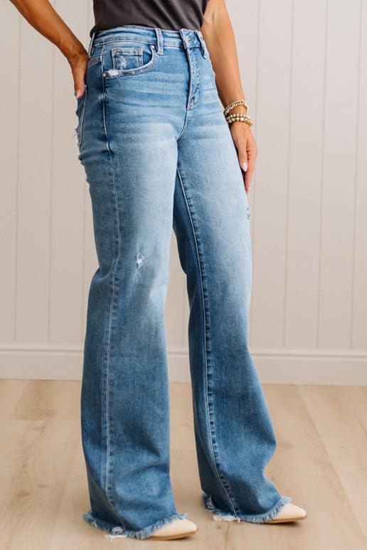 A person wearing light blue high-waisted flare jeans with a raw frayed hem, standing on a light wood floor against a white paneled wall.