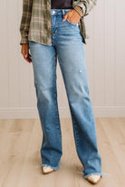 A person wearing light blue wide-leg denim jeans with a raw frayed hem, styled with a green plaid shirt and cream pointed-toe heels, standing in a room with light wood floors and white paneled walls.