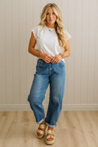Woman wearing a white top with lace sleeves and blue jeans standing against a light wooden paneled wall.