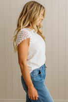 Woman wearing a white lace top and blue jeans against a beige wall.