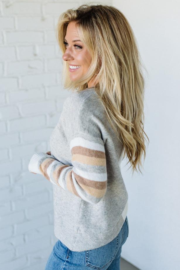 Woman wearing a gray sweater with striped sleeves against a white brick wall.
