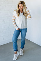 Woman wearing a striped sweater, jeans, and sneakers against a white brick wall.