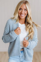 A smiling blonde woman wearing a light blue chunky cable-knit cardigan over a white t-shirt and light-wash jeans, standing against a neutral indoor background.