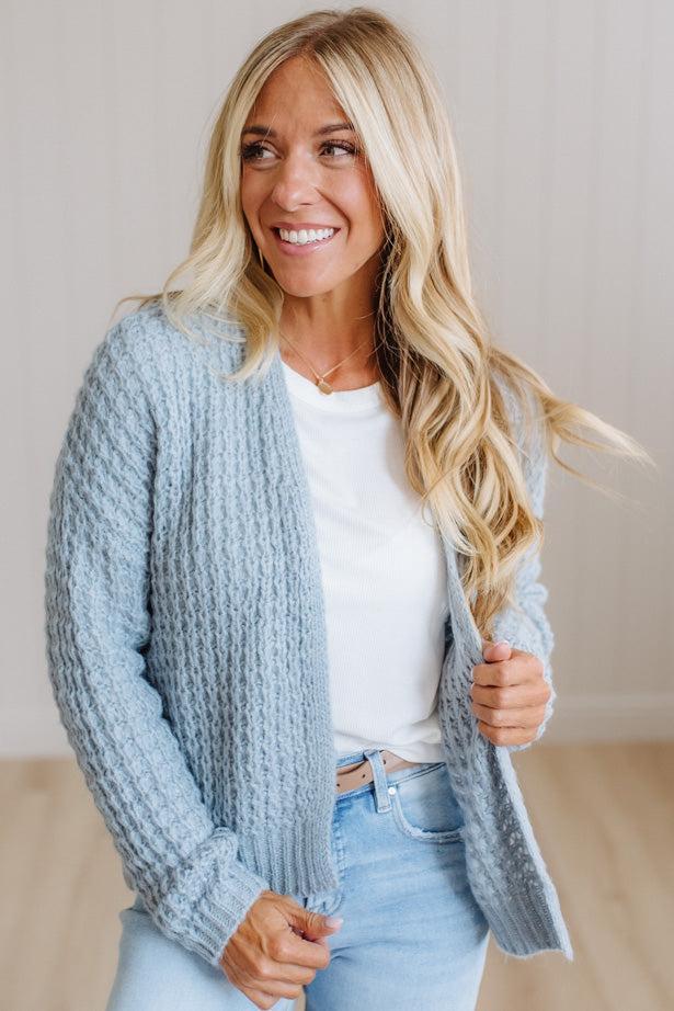 A smiling blonde woman wearing a light blue chunky cable-knit cardigan over a white t-shirt and light-wash jeans, standing in a brightly lit indoor setting.
